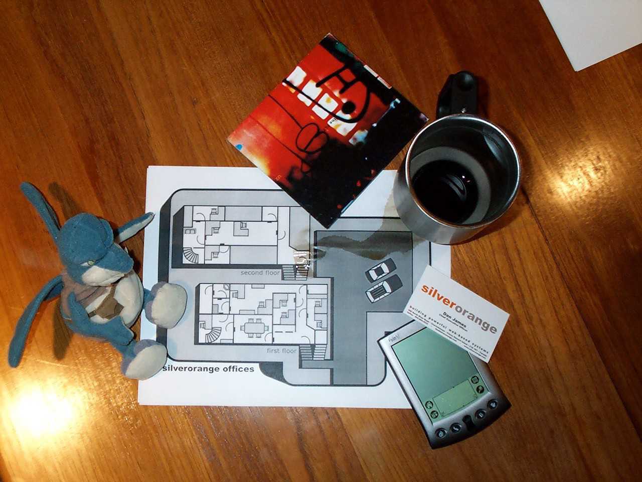 Pile of items on wooden table including Star Wars toy, CD case, coffee mug, business card, office map, and Palm Pilot