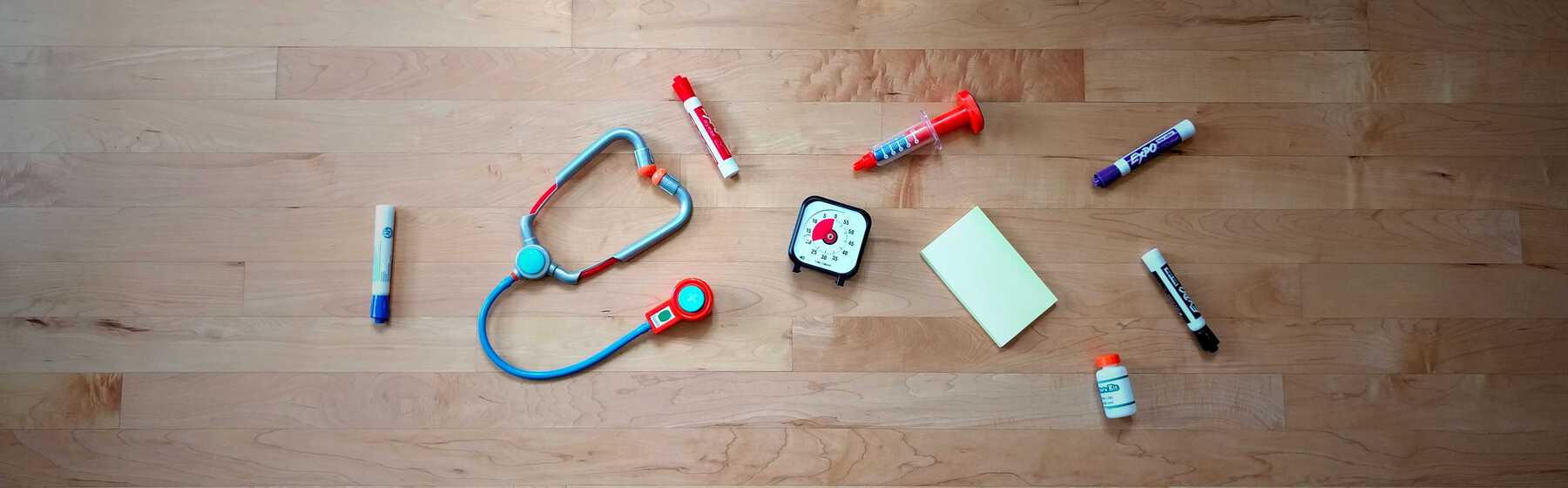 Whiteboard markers, post-it notes, a timer, and toy doctors tools arranged on a wood floor